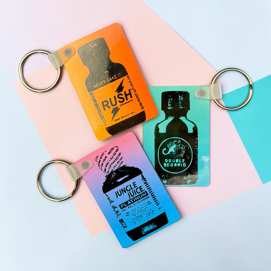 Colorful sublimated MDF keychains featuring poppers bottle designs arranged on pastel geometric background with metal keyrings attached.